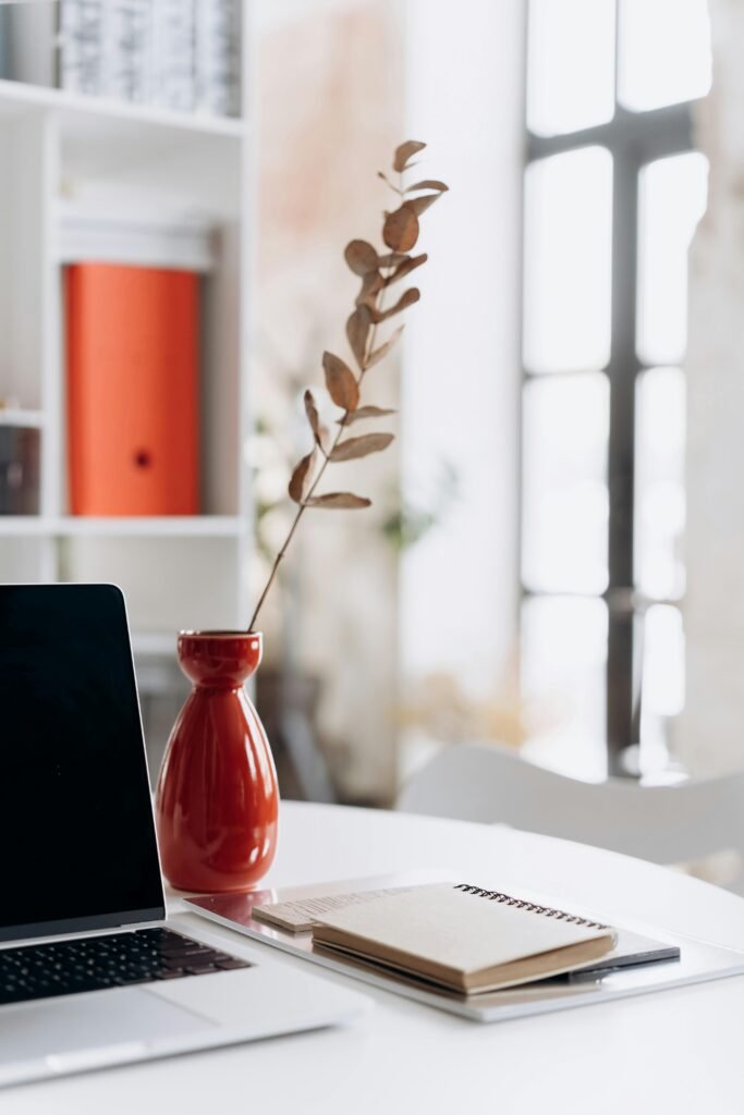 Modern workspace featuring a laptop, notebooks, and a red vase indoors with natural light.